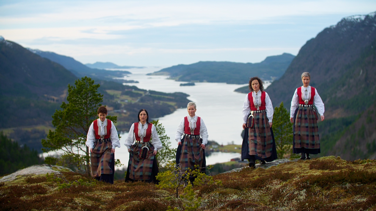 Fem kvinnor iklädda traditionell norsk bygdedräkt som går uppför ett berg.