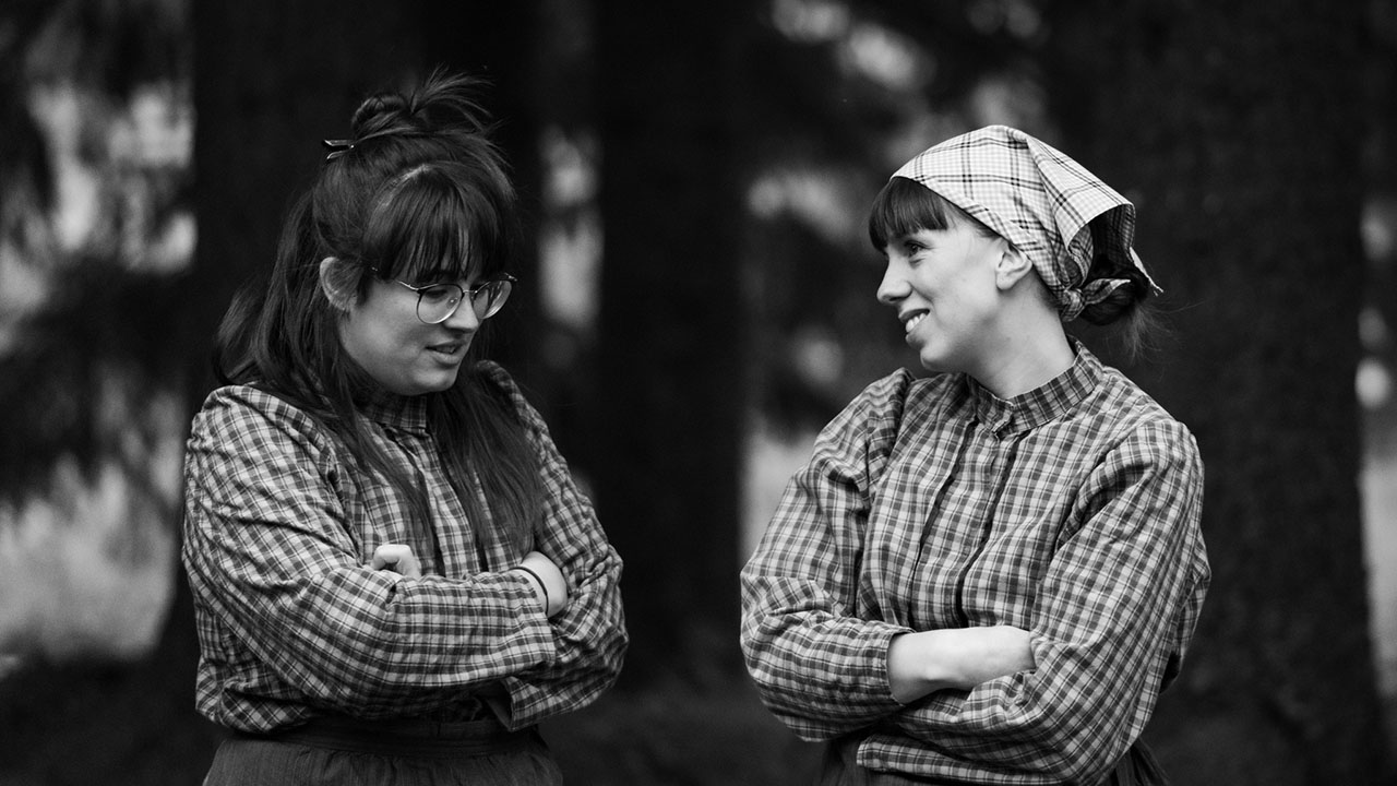 Svartvitt foto av Elsa och Sigrid utomhus. De bär rutiga blusar och Sigrid bär en hätta.